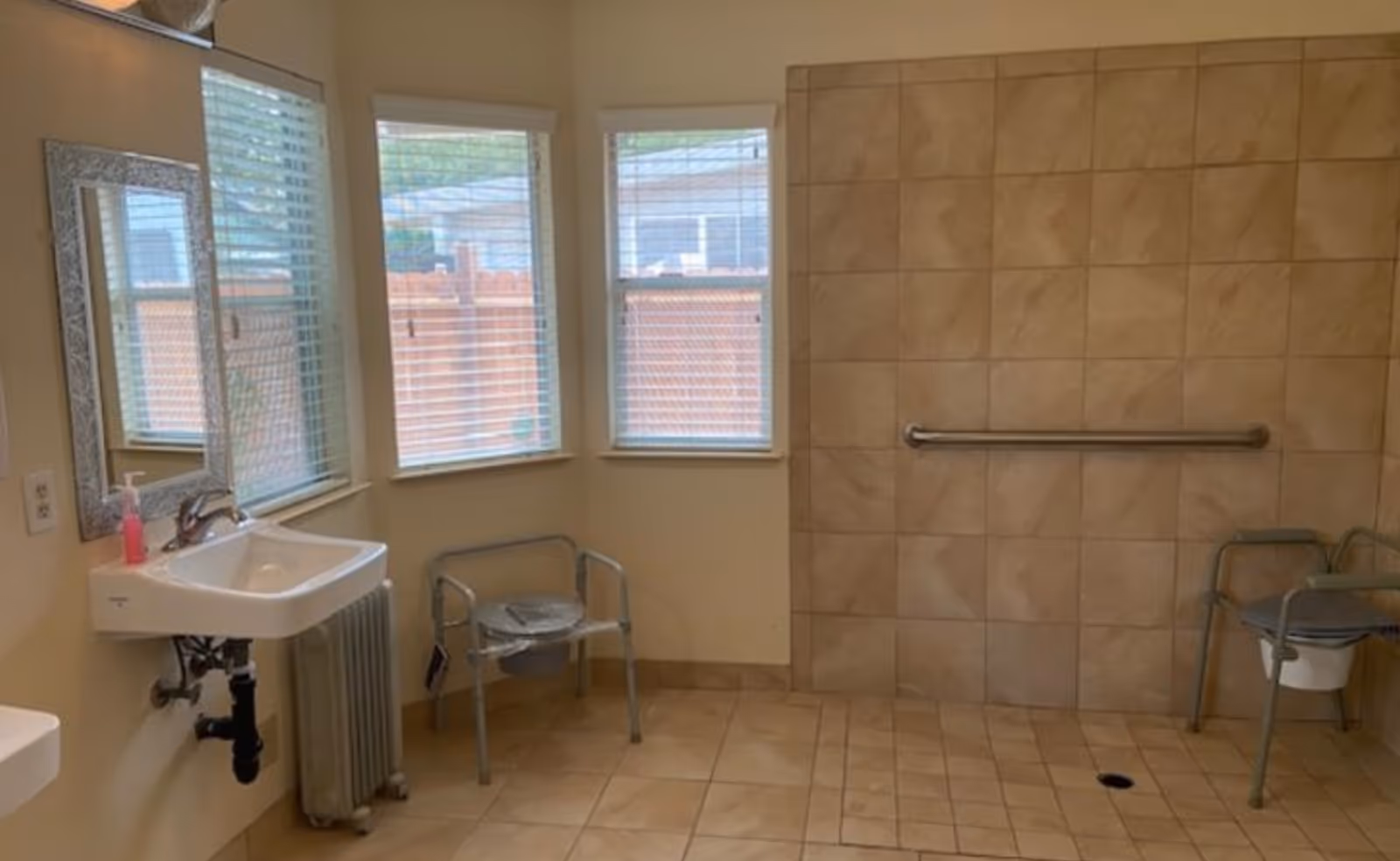 A spacious bathroom with beige tiled floor and walls, two windows with blinds, a wall-mounted sink with a mirror above it, a handrail on the tiled wall, and two metal commode chairs with plastic seats.