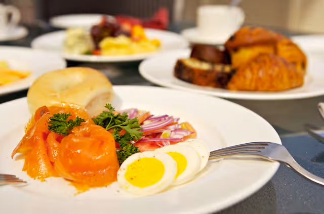 A close-up of a breakfast plate featuring smoked salmon garnished with parsley, sliced hard-boiled eggs, and red onion slices, with a bagel on the side. In the background, there are plates with croissants, bread, and other breakfast items along with white coffee cups.