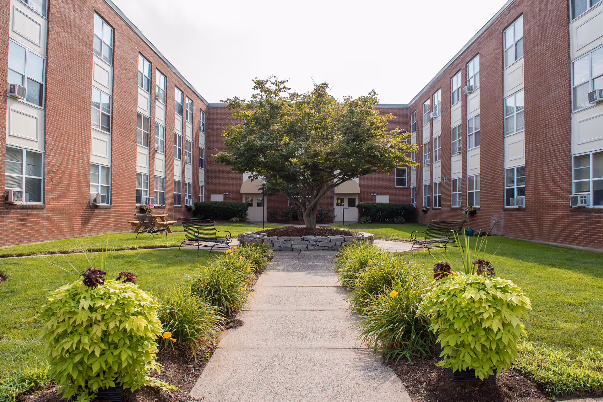 Well-maintained courtyard of a red-brick senior living building with a central tree, benches, walkways, and potted plants.