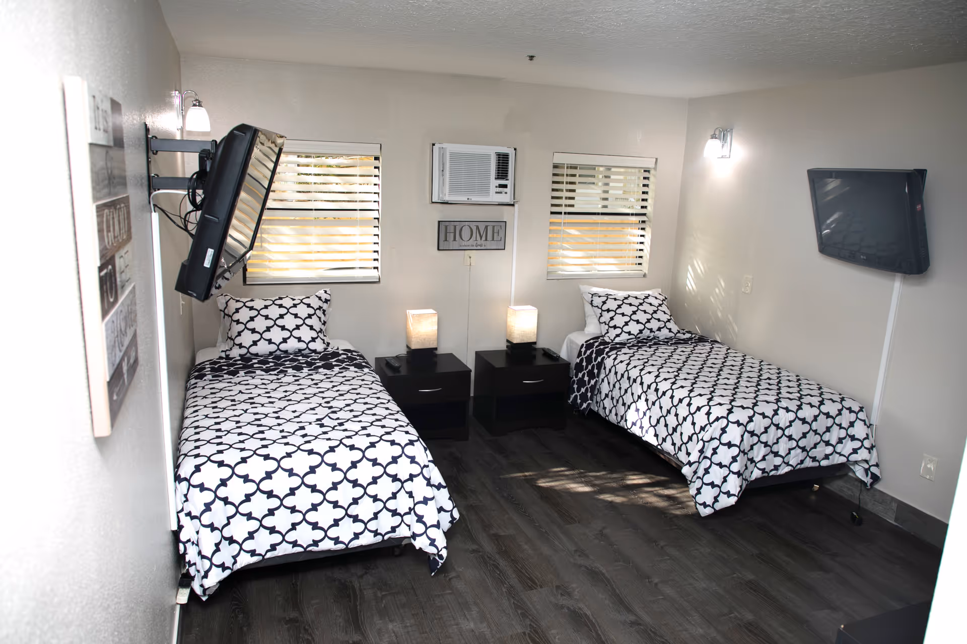 A bright bedroom with two single beds, each with black and white patterned bedding. Between the beds are two black nightstands with lamps. Two windows with blinds are on the wall behind the beds, along with a wall-mounted air conditioning unit and a decorative sign that reads 'HOME'. Two wall-mounted televisions are positioned on opposite walls.