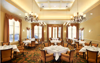A spacious dining room with multiple tables covered in white tablecloths, each set with napkins and silverware. The room features large windows with brown curtains, ornate chandeliers hanging from a high ceiling, and patterned carpet flooring. The walls are painted a warm beige color.