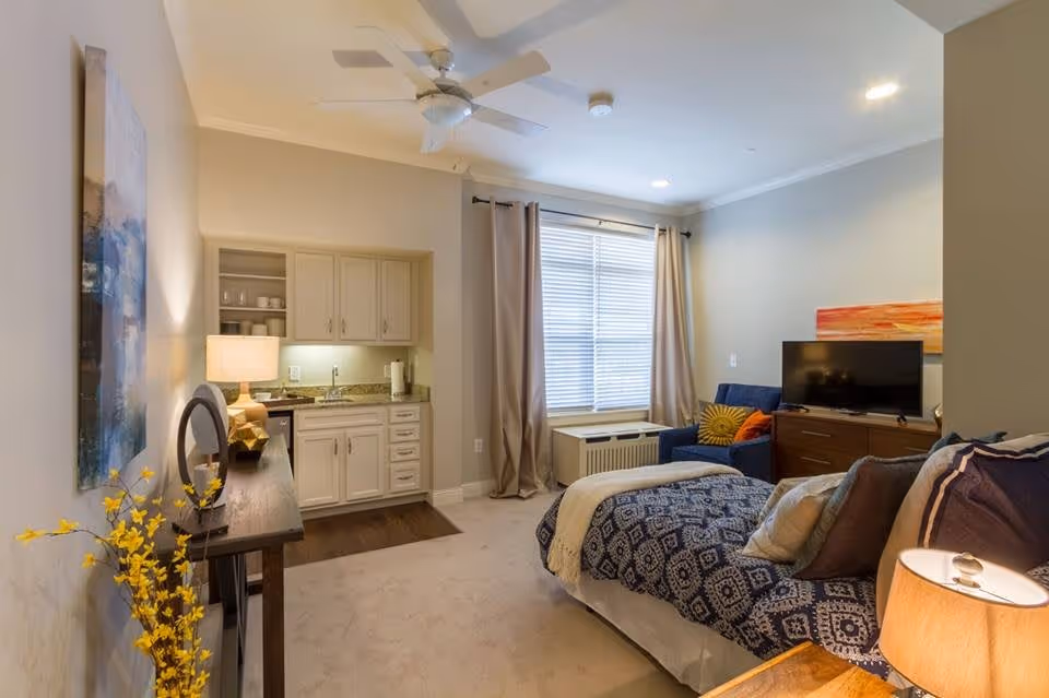 A cozy living space featuring a bed with patterned blue and white bedding, a wooden dresser with a flat-screen TV, a blue armchair with colorful pillows, a window with beige curtains, and a small kitchenette with cabinets, a sink, and a lamp on a console table with decorative items.