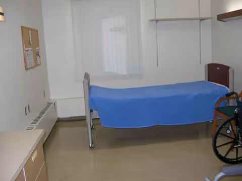 Hospital-style patient room with a bed, wheelchair, window, and storage cabinets.