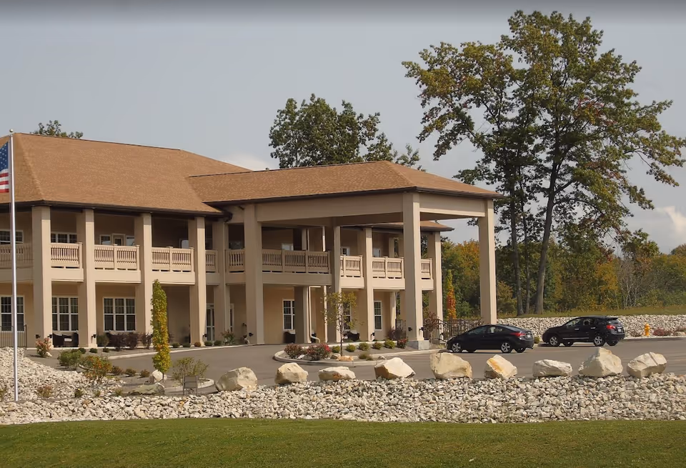 Front exterior of a two-story senior living building with a covered porte-cochere, columns, landscaped rock beds, and parked cars.