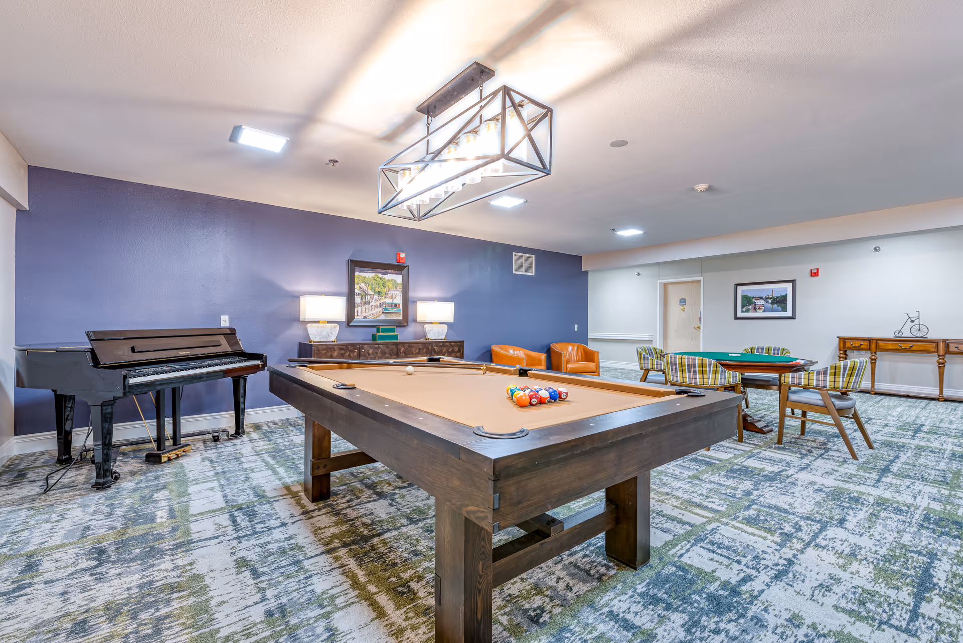 A recreational room with a pool table in the center, a black piano on the left, two orange armchairs against a blue wall, and a card table with four plaid chairs on the right. The room has patterned carpet flooring, framed artwork on the walls, and a rectangular ceiling light fixture above the pool table.
