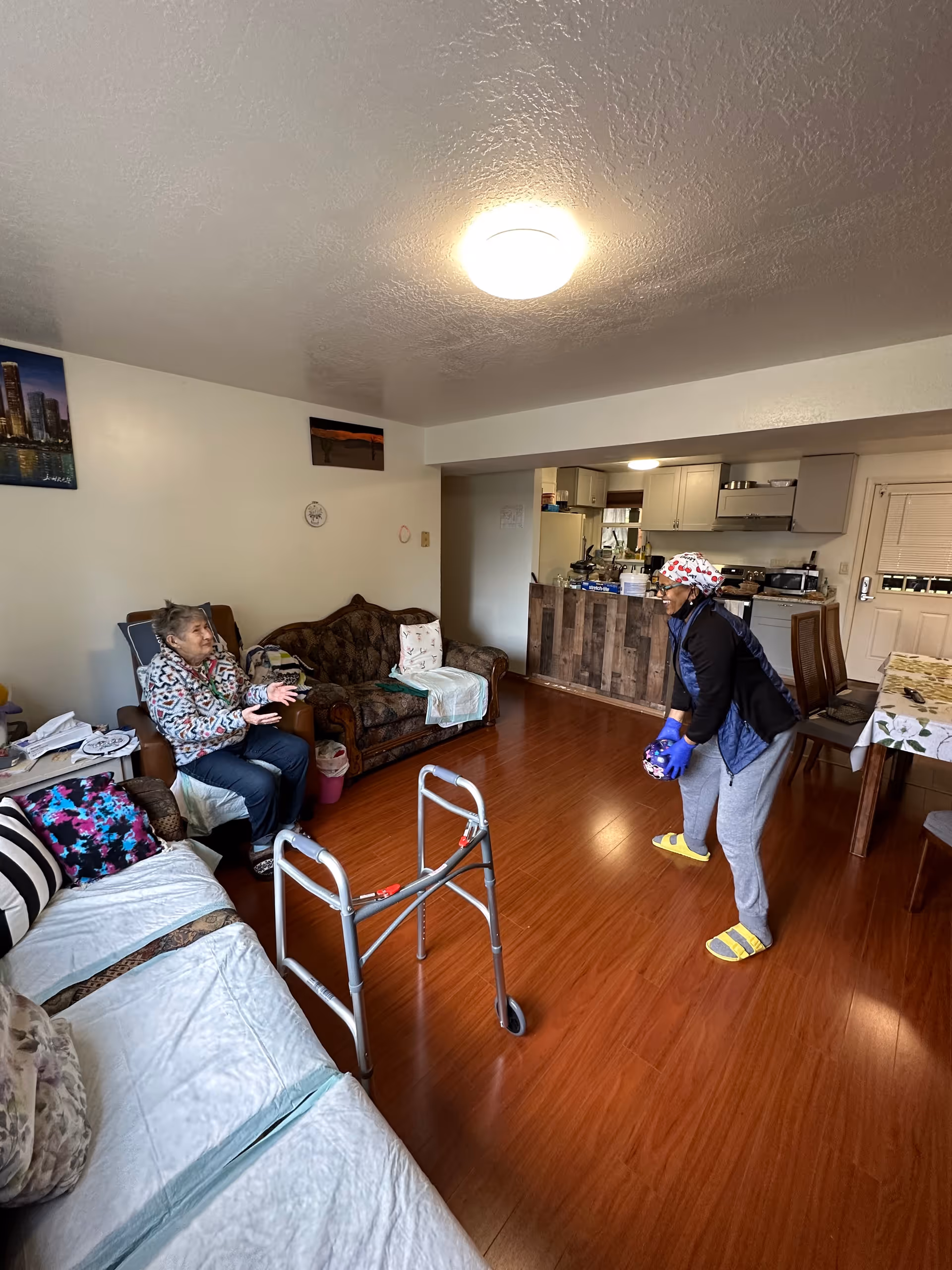 A cozy living room with wooden flooring featuring two elderly women interacting. One woman is seated on a brown armchair near a brown couch, while the other woman, wearing a headscarf and gloves, is standing and holding a ball. A walker is positioned in the middle of the room. The background shows a kitchen area with cabinets and appliances, and a dining table with chairs is visible on the right side.