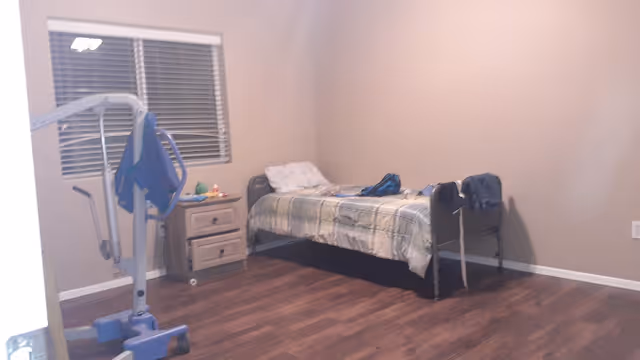 Sparsely furnished bedroom with a single bed, a nightstand under a window with blinds, and a medical lift on hardwood floors.
