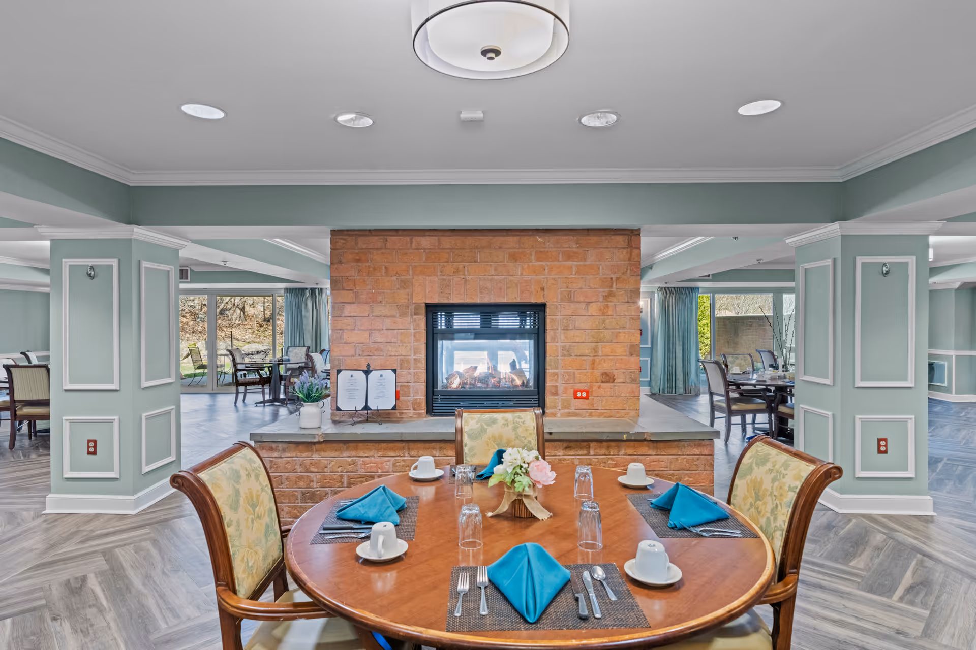 A dining area in a senior living facility with a round wooden table set for four people with blue napkins, white cups, and silverware. The table is positioned in front of a brick double-sided fireplace. The room has light green walls with white trim, large windows with blue curtains, and additional dining tables and chairs visible in the background.