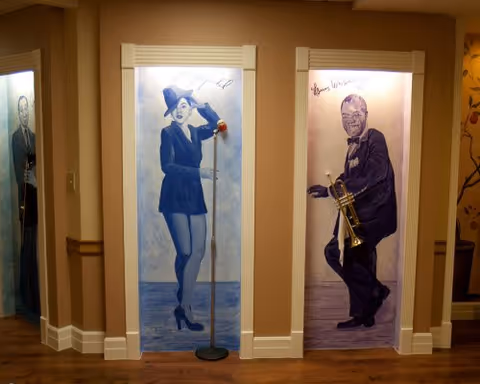 Three elevator doors in a hallway, each painted with a large mural of a person. The left door shows a man in a suit, the middle door features a woman in a blue outfit and hat holding a microphone stand, and the right door depicts a man in a tuxedo playing a trumpet. The walls are beige with white trim and the floor is wooden.
