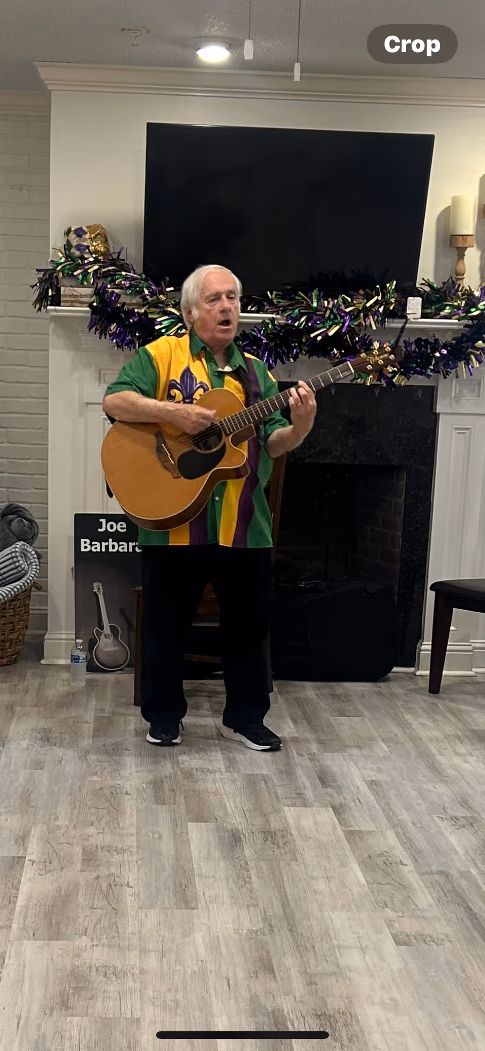 An elderly man wearing a colorful Mardi Gras-themed shirt is playing an acoustic guitar and singing in a room with a fireplace and a large TV mounted above it. The fireplace mantel is decorated with purple, green, and gold garlands. There is a basket with blankets on the left and a chair on the right.