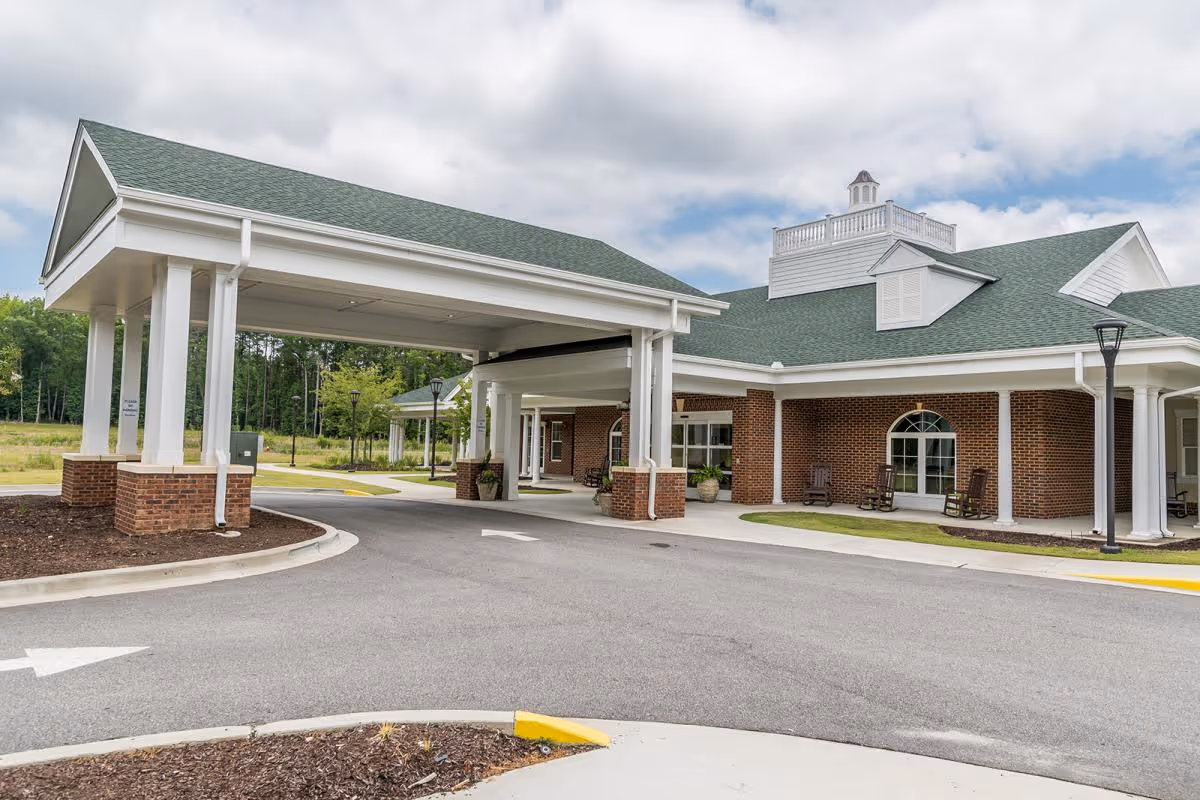 Exterior view of a senior living facility named Rice Estate featuring a covered entrance with white columns and brick base, green roof, and a paved driveway with directional arrows. The building has large windows and rocking chairs on the porch, with trees and grass in the background under a partly cloudy sky.