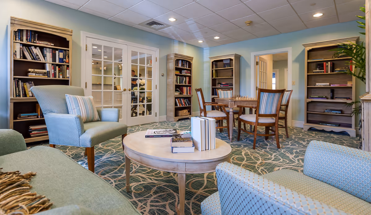 Comfortable common room with armchairs, a round coffee table, bookshelves and a table with chairs in a bright, carpeted lounge.