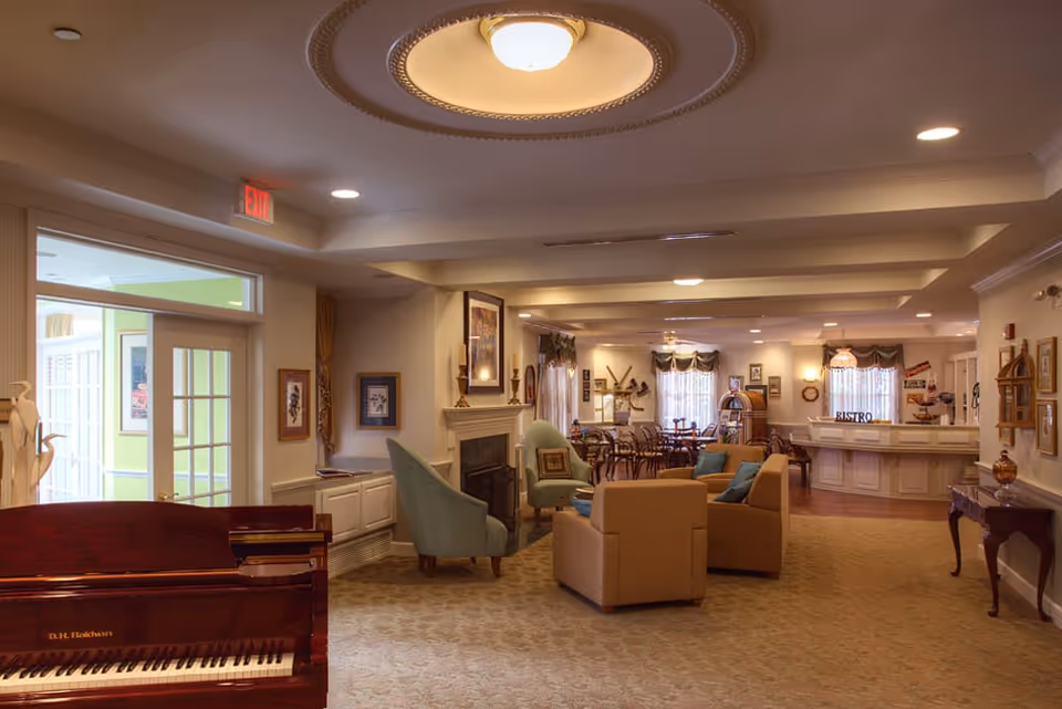 A spacious and warmly lit common area in a senior living facility featuring a piano in the foreground, comfortable seating with armchairs and sofas around a fireplace, and a dining area with tables and chairs in the background. The room has decorative ceiling molding, framed artwork on the walls, and a bistro counter at the far end.