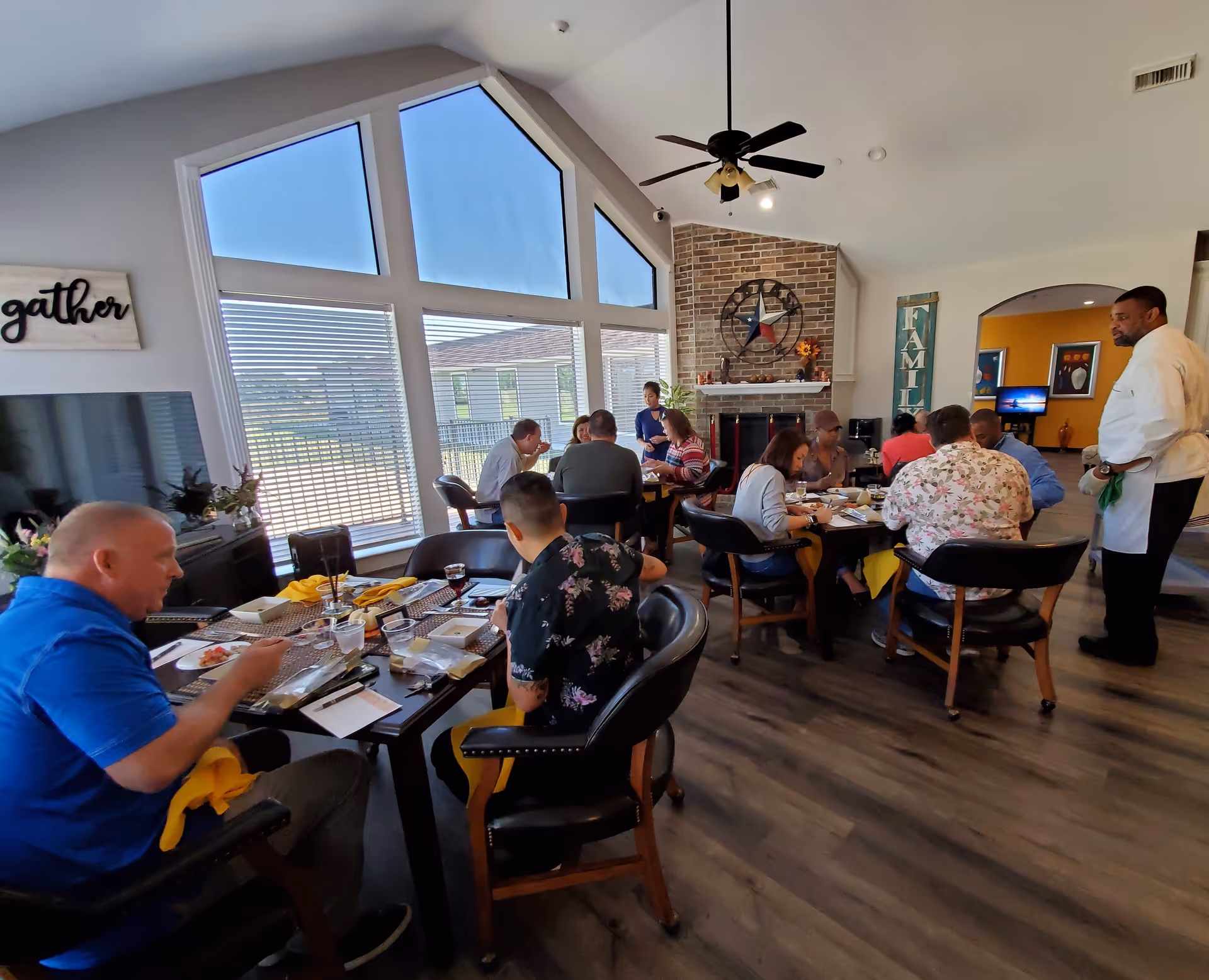 A group of people seated around two dining tables inside a residential care facility, enjoying a meal together. The room features large triangular windows, a brick fireplace with a Texas-themed clock above it, and wall decorations including signs that say 'gather' and 'family'. A man in a white chef's coat stands nearby, observing the diners.