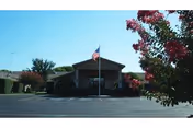 Front exterior view of a single-story building with an American flag on a flagpole in front, surrounded by trees and bushes under a clear blue sky.