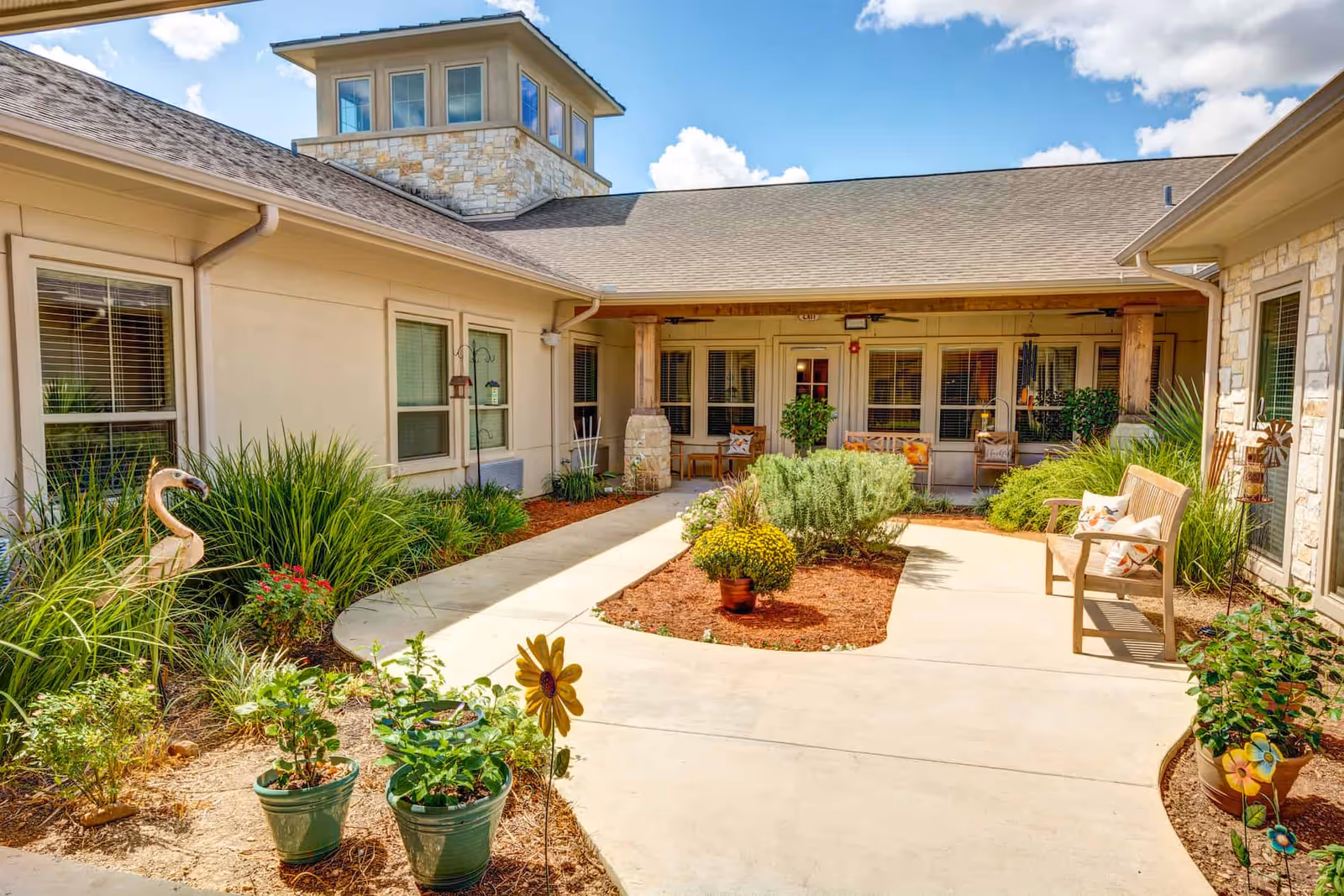 Outdoor courtyard area at The Auberge at Cedar Park featuring a paved walkway surrounded by various plants and flowers in pots and garden beds. There are wooden benches with decorative pillows, a flamingo garden statue, and a building with multiple windows and a stone tower under a partly cloudy blue sky.