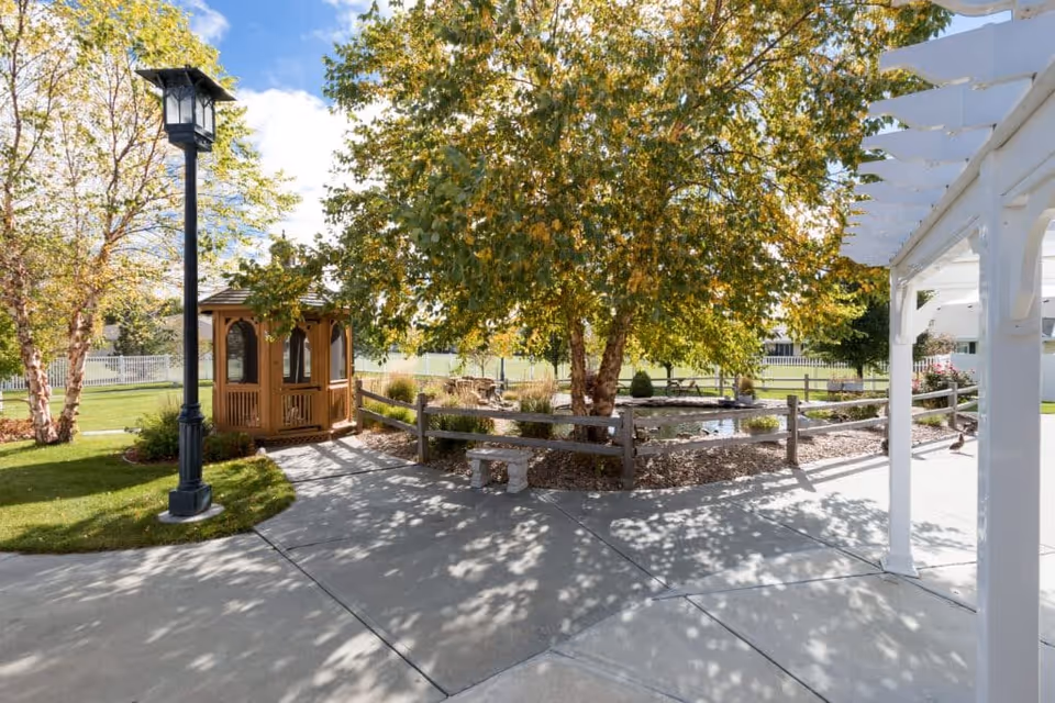 Outdoor garden area with a wooden gazebo, a black lamp post, a tree with green leaves, a small pond surrounded by a wooden fence, a stone bench, and a white pergola structure on the right side under a blue sky with some clouds.