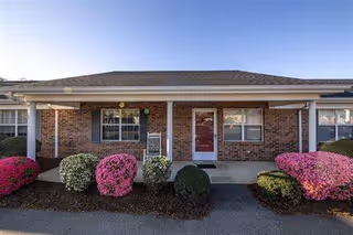 Single-story brick retirement cottage front with a covered porch, white columns, a red door, and pink flowering shrubs.