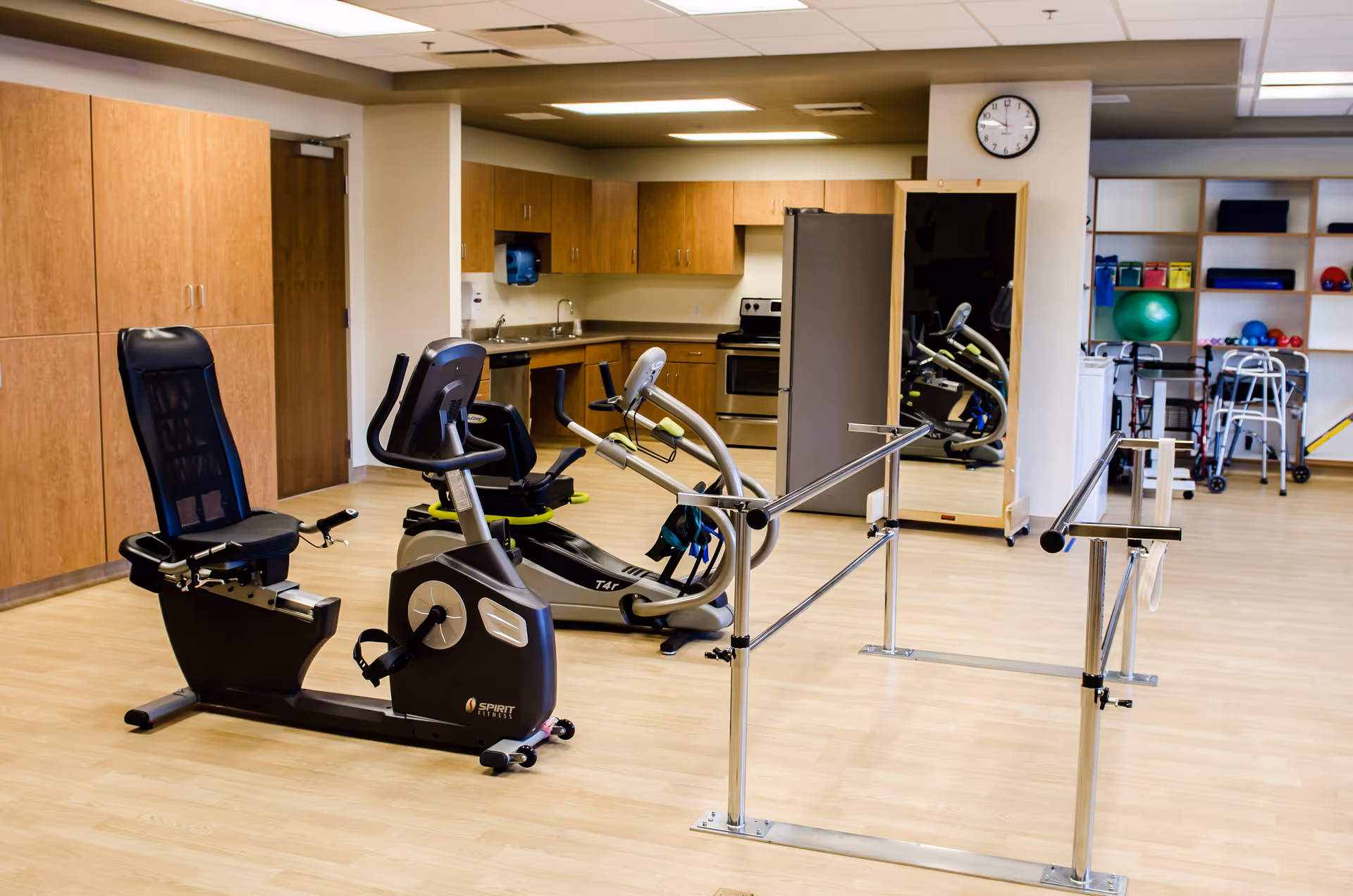 A spacious rehabilitation room with exercise equipment including a recumbent bike and an elliptical machine, parallel bars for walking practice, a large mirror, wooden cabinets, and shelves with exercise balls and walkers in the background.