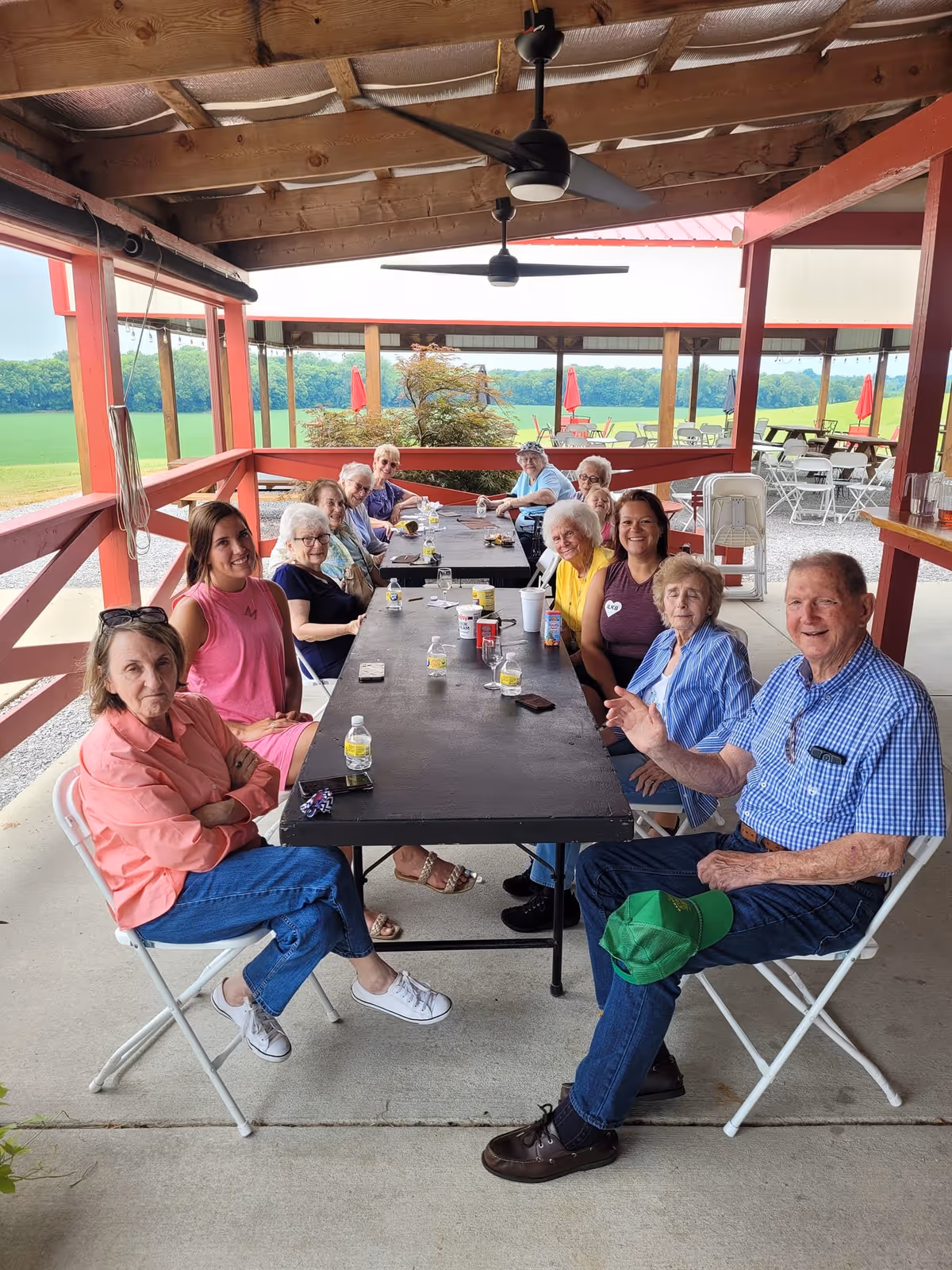 A group of elderly people and a few younger adults sitting around a long black table under a covered outdoor patio. They are smiling and appear to be enjoying a social gathering. The patio has a wooden roof with ceiling fans, and there is a green field visible in the background along with red umbrellas and outdoor seating.