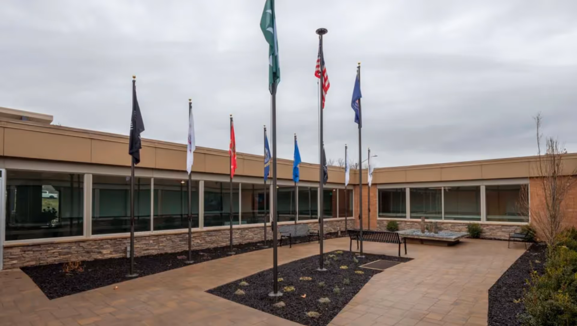 Outdoor courtyard area at Heritage Park Healthcare and Rehabilitation with multiple flagpoles displaying various flags, surrounded by a paved walkway, benches, and landscaping beds with mulch and small plants. The building exterior features large windows and a beige and stone facade under a cloudy sky.