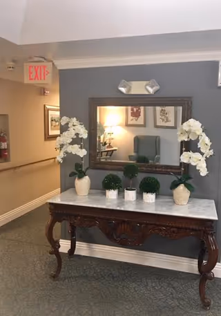 A hallway corner in an assisted living facility featuring a decorative wooden table with a white marble top. On the table are two vases with white orchids and three small potted green plants. Above the table is a large rectangular mirror reflecting a lamp, framed artwork, and a green armchair. An exit sign and fire extinguisher are visible on the left side of the hallway.