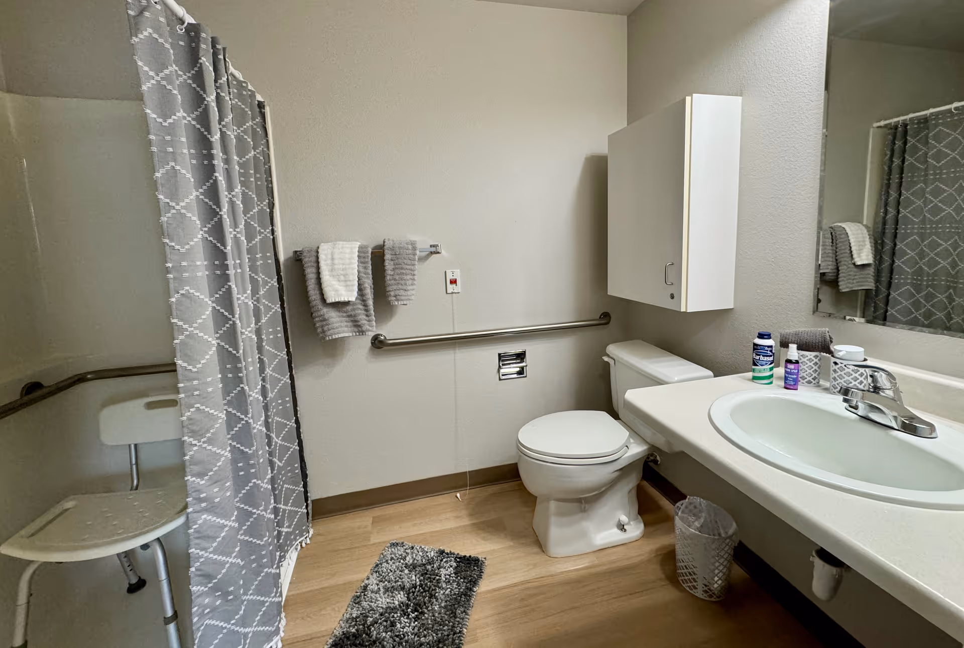 A senior living facility bathroom featuring a white toilet, a sink with a countertop, a large mirror, and a wall-mounted cabinet. There is a gray and white patterned shower curtain partially covering a shower area with a white shower chair. Two towels hang on a towel rack above a grab bar on the wall. The floor is wood-style, and a small gray rug is placed in front of the shower.