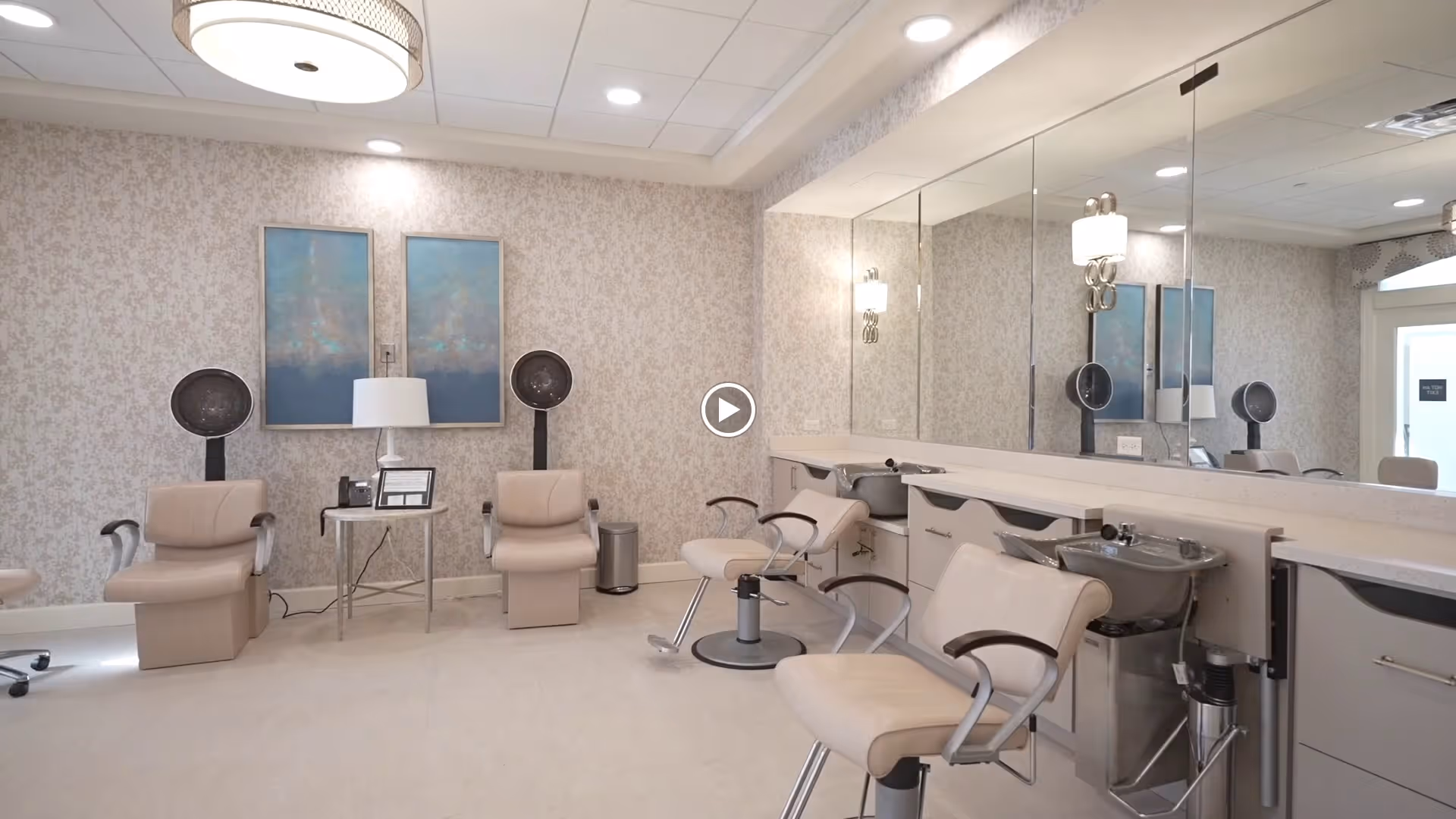 A modern hair salon area with beige salon chairs, hair dryers, and wash basins. The room has a large mirror along one wall, two abstract paintings on the opposite wall, and soft lighting from ceiling fixtures. The decor is light and neutral with patterned wallpaper.