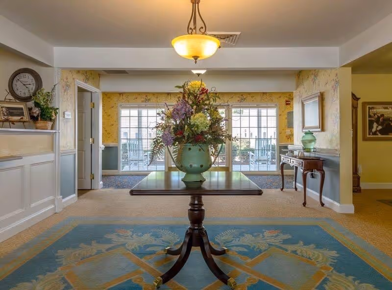 Bright assisted-living lobby with a central round table holding a large floral arrangement, patterned rug, and glass doors letting in natural light.