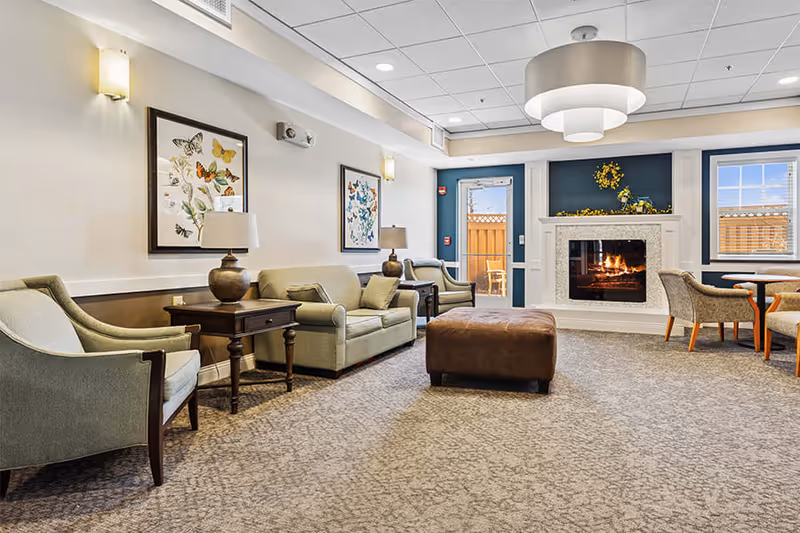 A cozy living room area with beige and green upholstered chairs and sofas, a brown ottoman in the center, two framed butterfly artworks on the wall, table lamps on wooden side tables, a lit fireplace with a white mantel decorated with yellow flowers, and a round table with chairs near windows and a glass door leading outside.