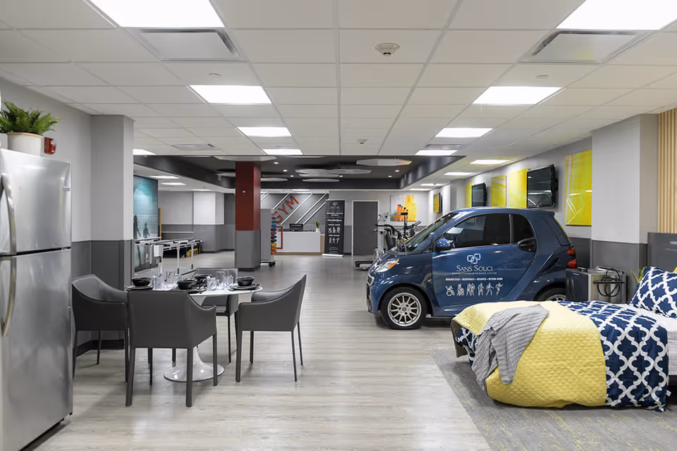 Interior view of a multi-purpose room in Sans Souci Rehabilitation and Nursing Center featuring a small dining table with four chairs, a stainless steel refrigerator, a bed with blue and yellow bedding, a small blue car with the facility's logo, and exercise equipment in the background under bright ceiling lights.