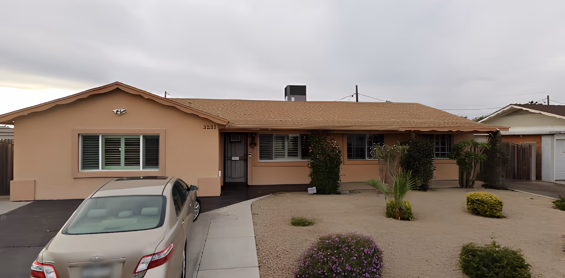 Front exterior view of a single-story peach-colored house with a brown roof, a driveway with a parked beige car, and a front yard with desert landscaping including small bushes and plants.