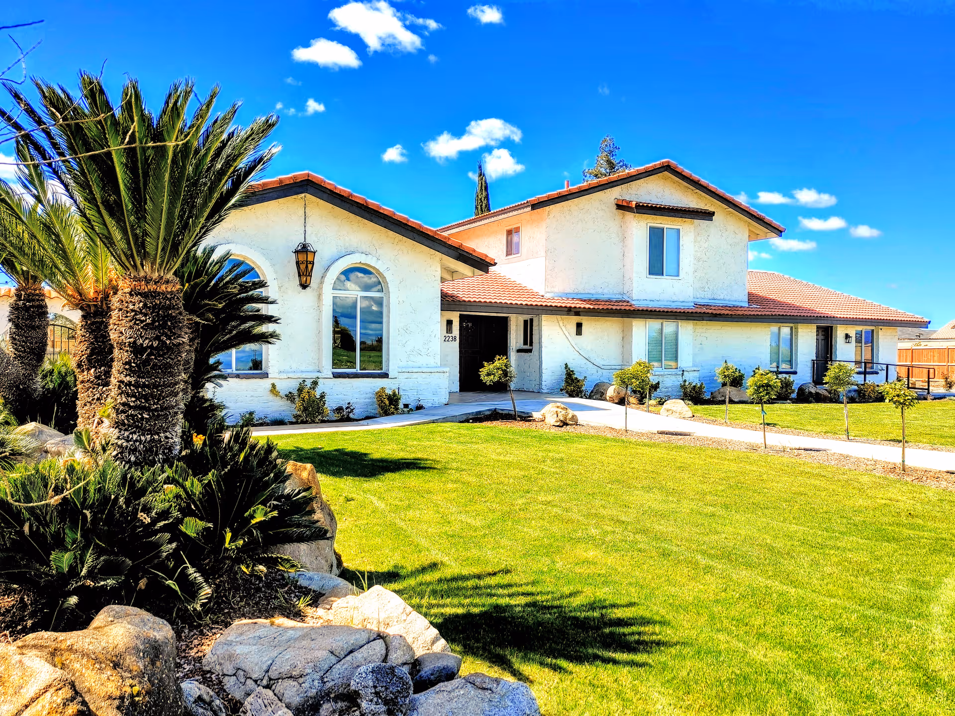 A bright, sunny exterior view of a two-story residential care facility with white walls and a red-tiled roof. The building is surrounded by a well-maintained green lawn, small trees, and palm plants. The sky is clear with a few scattered clouds.