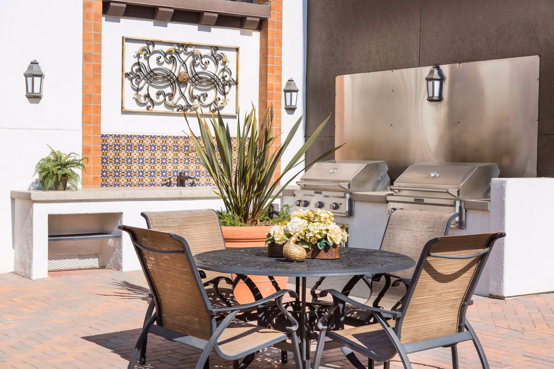 Outdoor patio area with a round metal table and four chairs. The table has a decorative centerpiece with flowers and a small golden bird figurine. Behind the table are two stainless steel grills mounted on a white wall with a decorative tile backsplash and ornamental wrought iron wall art. There are also two wall-mounted lantern-style lights and a large potted plant.