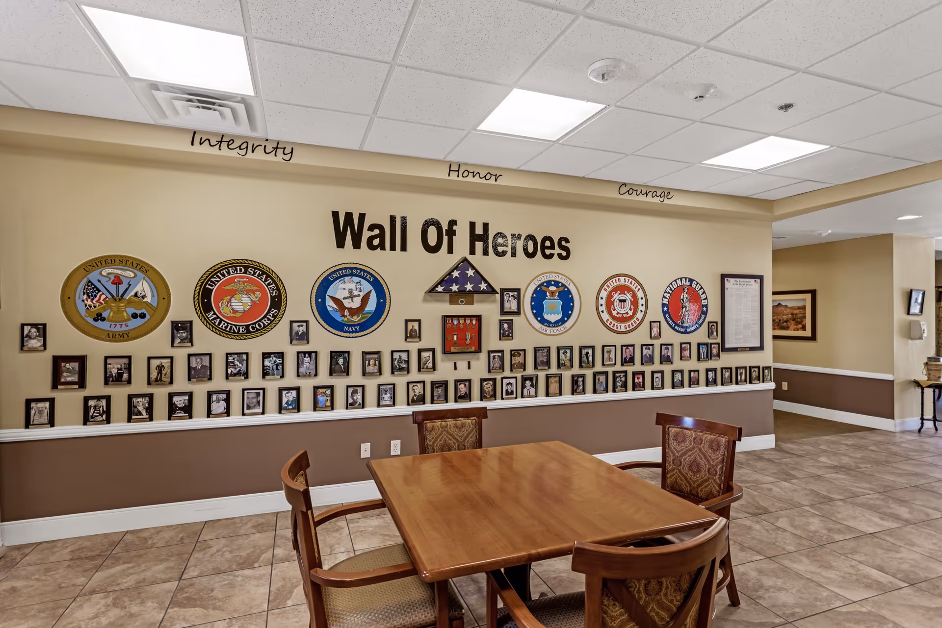 Interior view of a room with a wooden table and four chairs in front of a wall display titled 'Wall Of Heroes' featuring emblems of United States Army, Marine Corps, Navy, Air Force, Coast Guard, and National Guard, along with numerous framed photographs and a folded American flag. Words 'Integrity', 'Honor', and 'Courage' are written above the display.