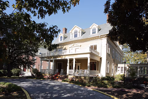 Exterior view of a large, light-colored, multi-story assisted living community building with a covered porch, surrounded by trees and landscaping under a clear blue sky.