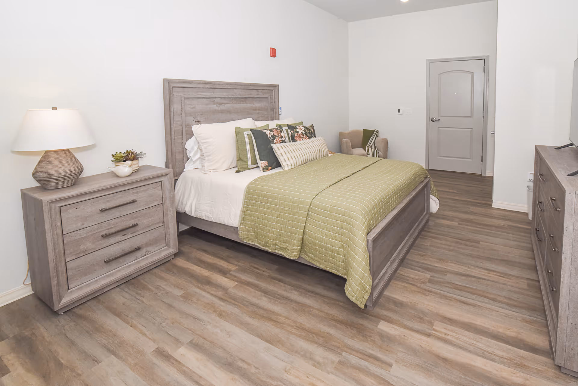A bedroom with a wooden bed frame and headboard, neatly made with white sheets, green quilt, and multiple decorative pillows. To the left of the bed is a wooden nightstand with a lamp and a small plant. In the corner is a beige armchair with a green and white throw blanket. On the right side of the room is a wooden dresser with a flat-screen TV on top. The floor is wood laminate, and the walls are painted white.