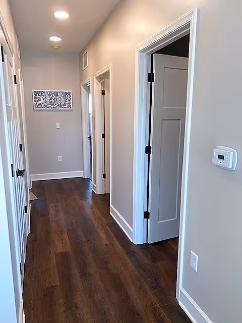 A hallway with dark wooden flooring and light gray walls. There are several white doors on both sides of the hallway, one of which is partially open. A thermostat is mounted on the right wall, and a framed artwork is hung at the end of the hallway. The ceiling has recessed lighting.