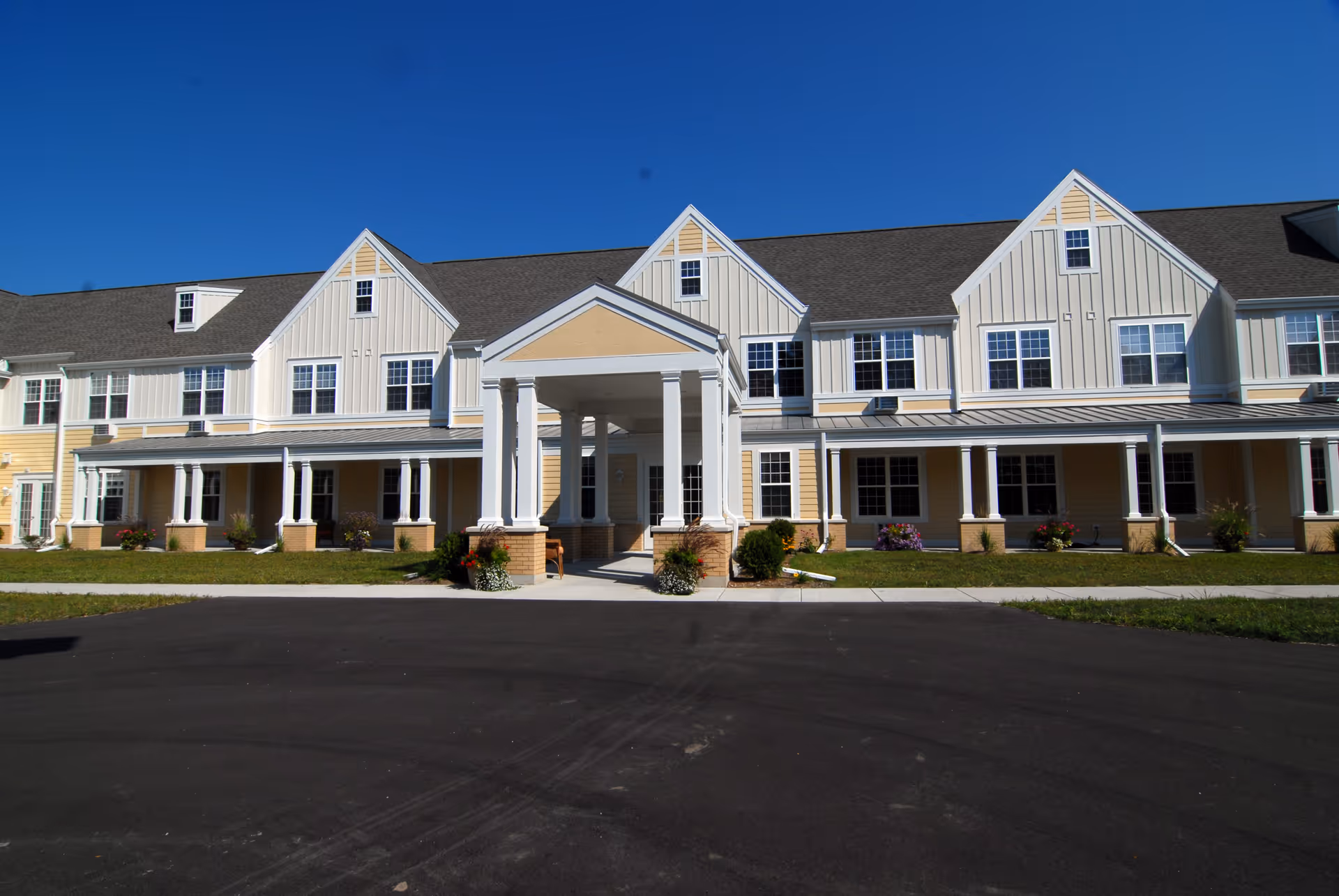Exterior front view of a two-story senior living facility building with a covered entrance supported by white columns, beige and white siding, multiple windows, and a clear blue sky.