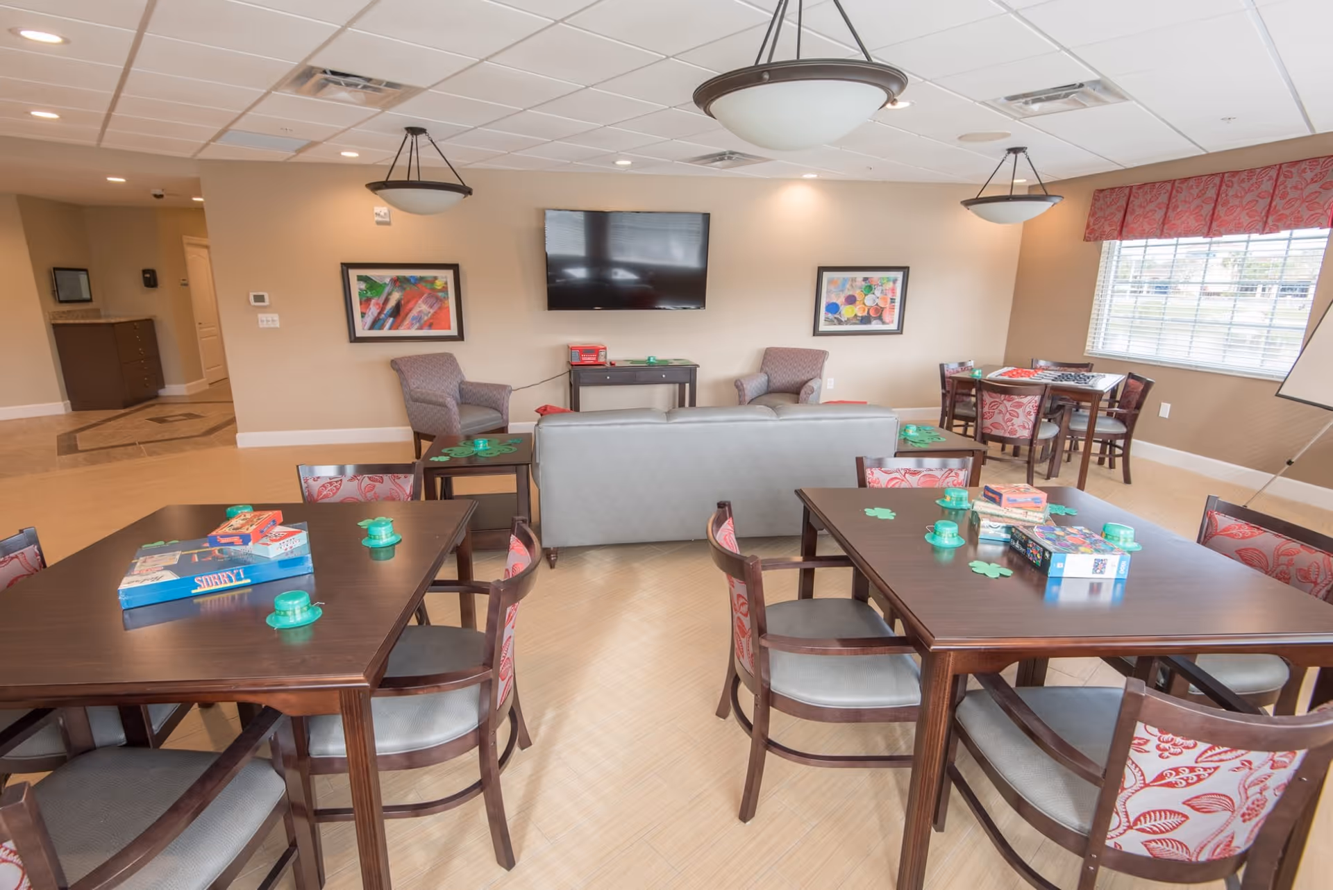 A bright senior living common room with tables and chairs set up for games, a sofa and armchairs facing a wall-mounted TV, and large windows.