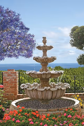 A multi-tiered stone water fountain surrounded by colorful flowers and greenery, with a brick wall and metal railing in the background. Purple and green trees frame the scene under a partly cloudy blue sky.