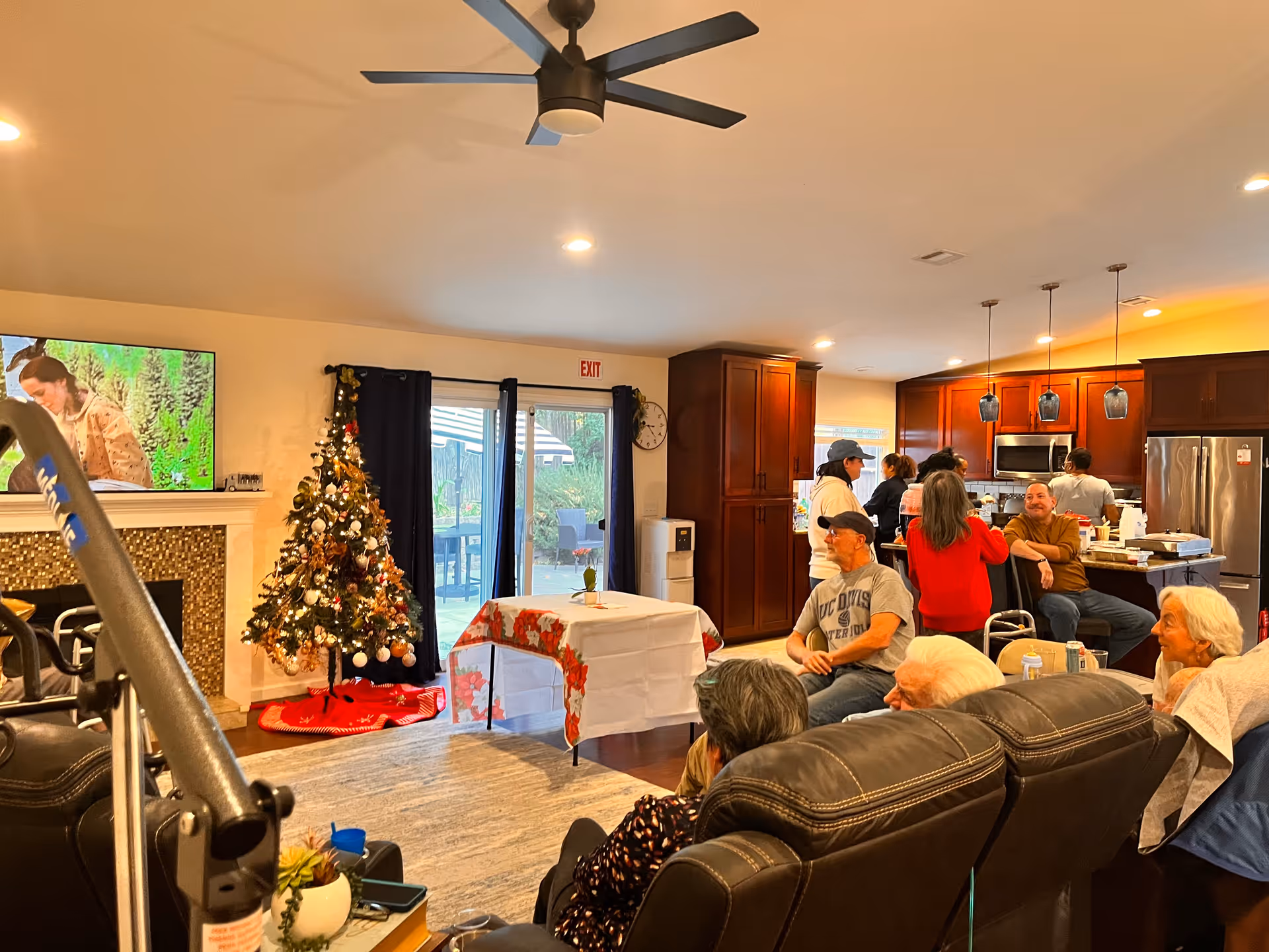 A cozy living room and kitchen area decorated for Christmas with a Christmas tree near a fireplace. Several people are sitting on leather chairs and standing near the kitchen island, engaging in conversation. The kitchen has wooden cabinets, stainless steel appliances, and pendant lights. A ceiling fan is visible above, and a TV is mounted above the fireplace.