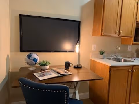 A small workspace area with a wooden desk, a blue upholstered chair, a globe, a small potted plant, an open book, a black mug, and a modern desk lamp. Mounted on the wall above the desk is a flat-screen television. To the right, there is a kitchenette with a sink, countertop, and wooden cabinets.