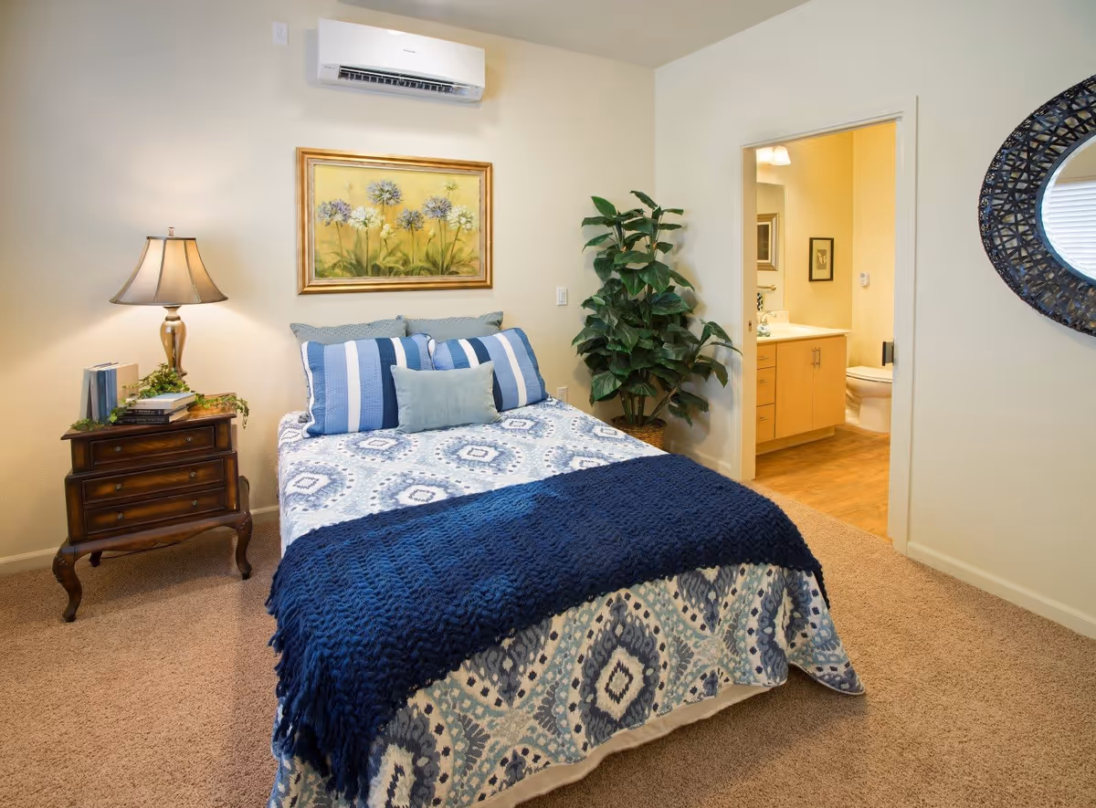 A cozy bedroom with a bed covered in a blue and white patterned quilt and blue pillows. There is a wooden nightstand with a lamp, books, and a small plant on the left side of the bed. A framed floral painting hangs on the wall above the bed. To the right, there is a doorway leading to a bathroom with a visible toilet and sink. A large green potted plant is placed near the bathroom entrance, and a decorative round mirror is mounted on the wall to the right.