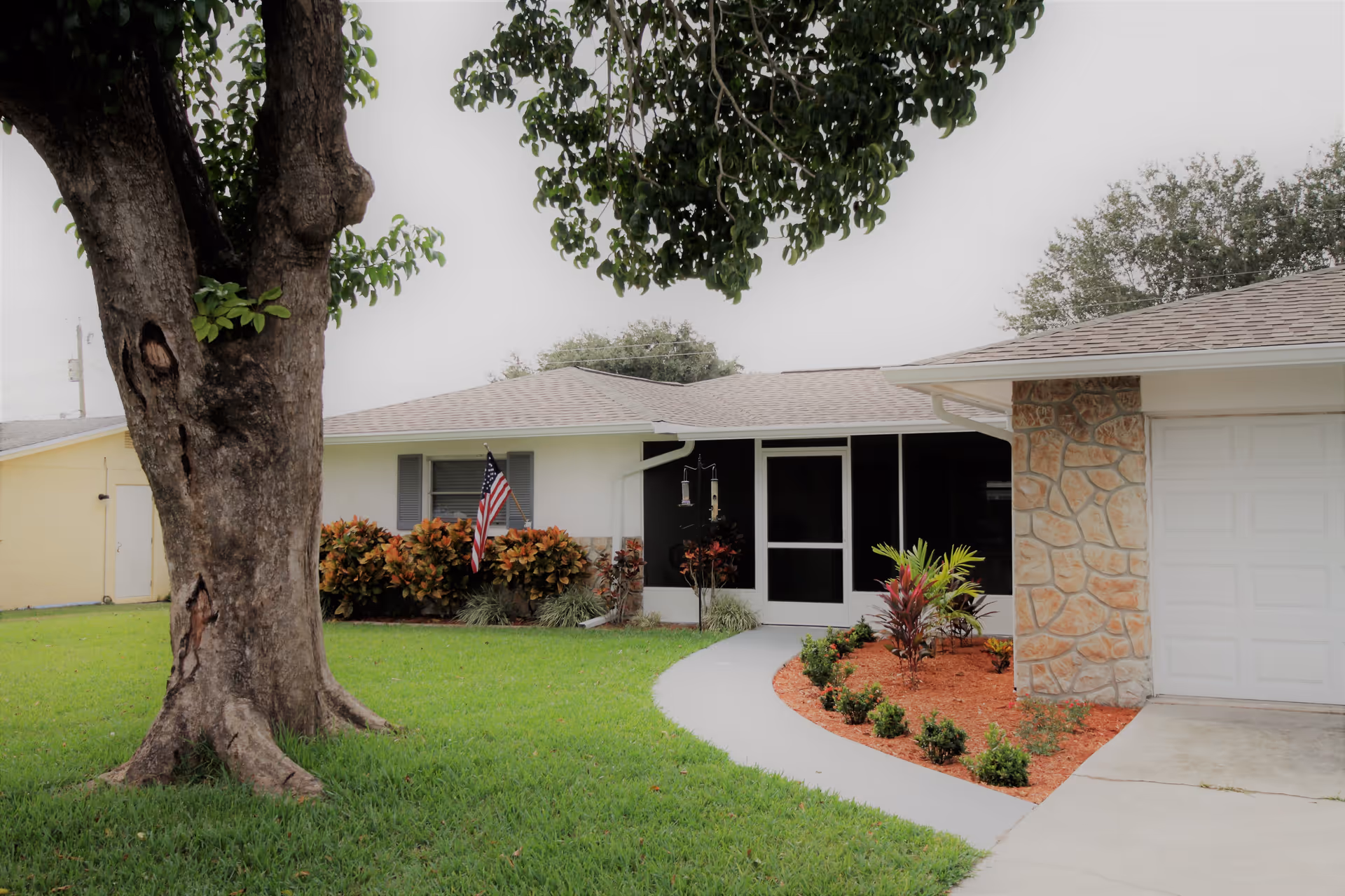 Front exterior view of a single-story assisted living facility building with a large tree in the grassy front yard, a curved concrete walkway leading to a screened porch entrance, landscaped plants with mulch, and an American flag near the window.