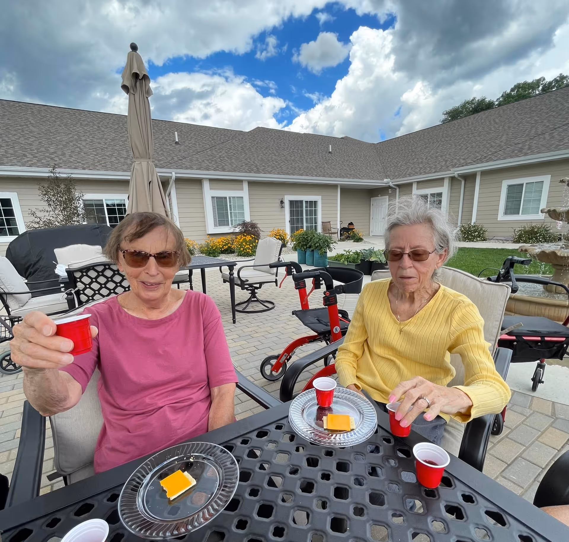 Two elderly women sitting outside at a black metal table with small red cups and plates with cheese and crackers. They are in a courtyard area with patio furniture, a closed umbrella, and a building with beige siding and white trim in the background. One woman is wearing a pink shirt and sunglasses, holding up a red cup, and the other woman is wearing a yellow sweater and glasses, reaching for a red cup. There are walkers behind them and a partly cloudy blue sky above.