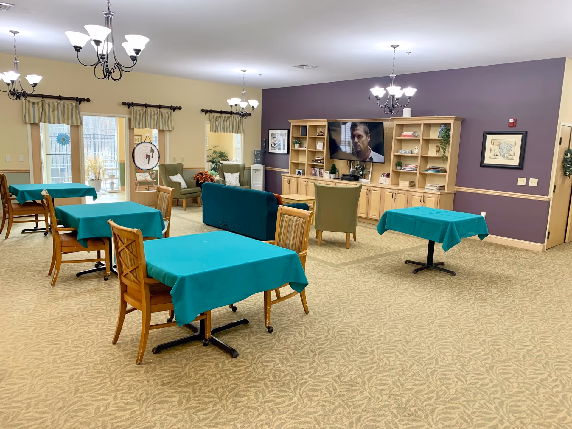 Senior living lobby with teal-draped tables, chairs, and a TV mounted on a purple accent wall.