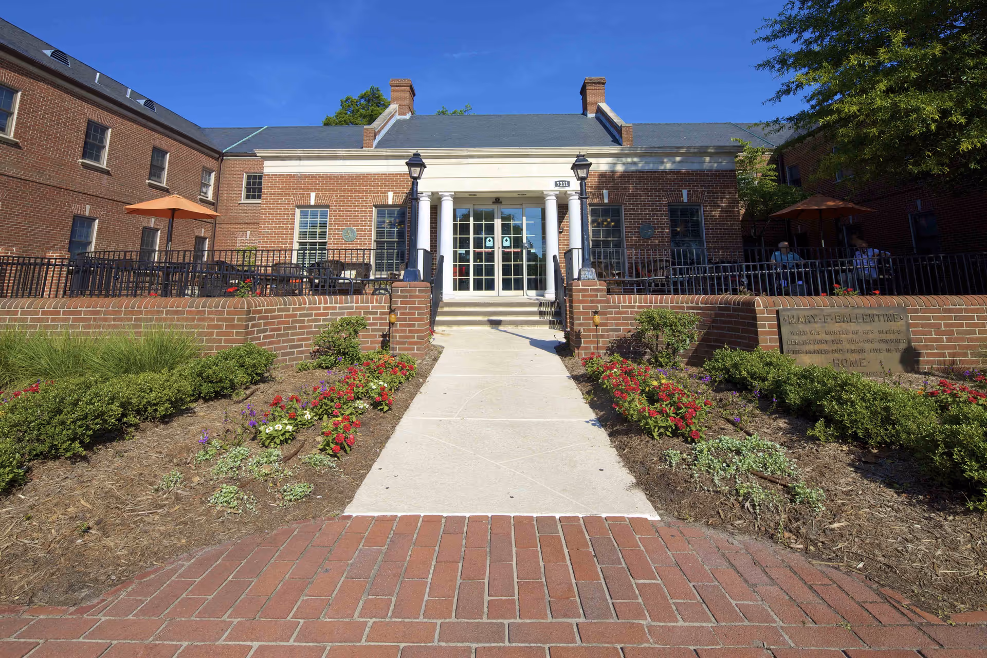 Front exterior view of Commonwealth Senior Living at the Ballentine building with a brick facade, a central entrance with white columns, outdoor seating areas with umbrellas, and landscaped flower beds along the walkway leading to the entrance.