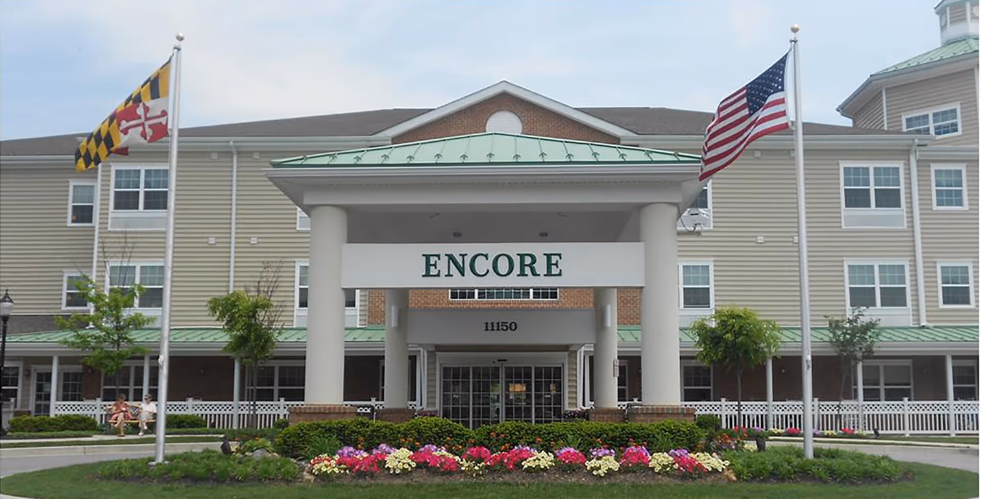 Front exterior view of the Lorien Encore senior living facility with a covered entrance labeled 'ENCORE'. The building is three stories tall with beige siding and white trim. Two flagpoles stand in front, one with the Maryland state flag and the other with the American flag. There are colorful flower beds and neatly trimmed bushes in front of the entrance. Two people are sitting on a bench to the left side of the entrance.