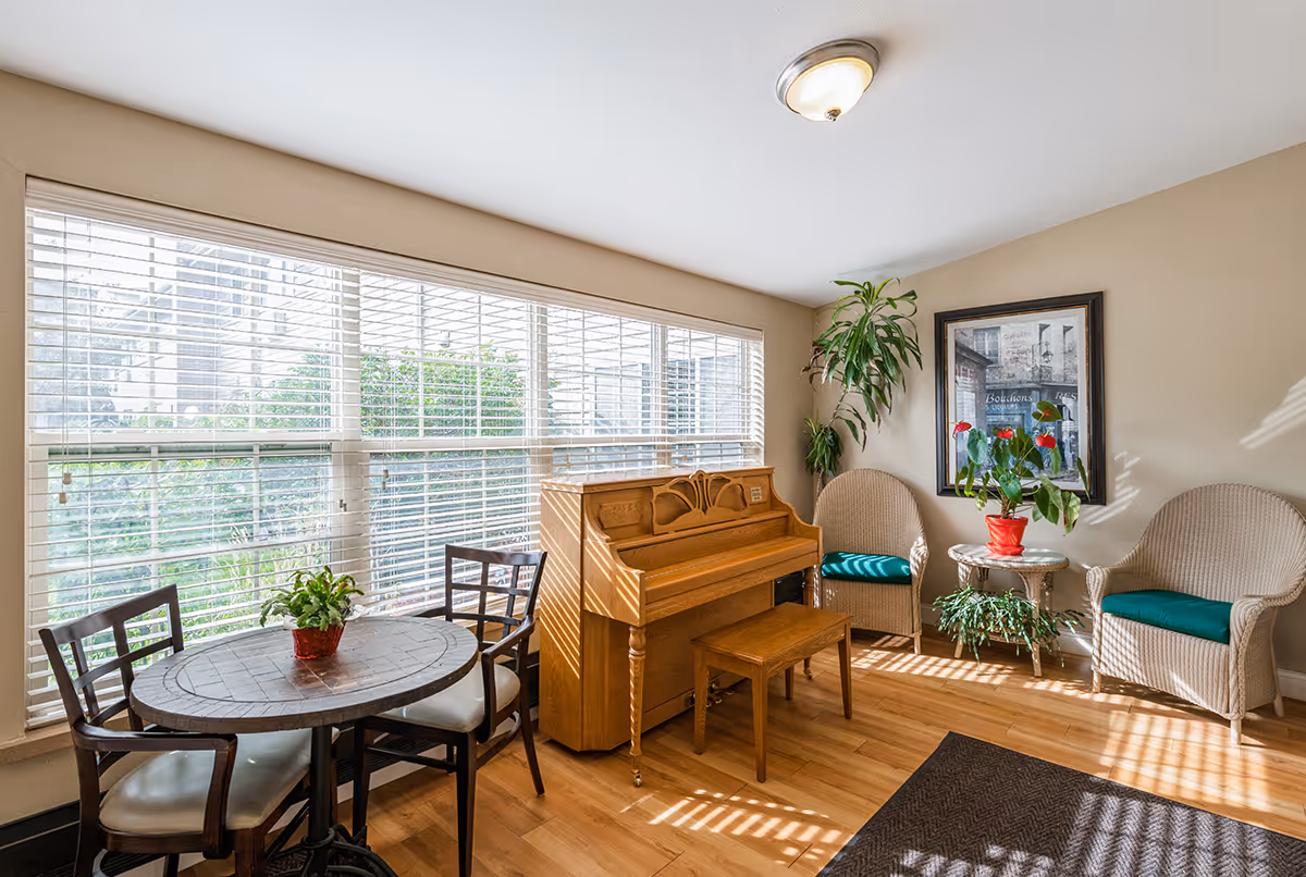 A bright room with large windows covered by white blinds, featuring a wooden piano with a matching bench, a round table with two chairs, two wicker armchairs with green cushions, a small round side table with a red potted plant, and additional green plants in the corners. The floor is wooden and the walls are painted beige.